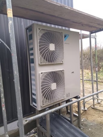 Zewnętrzna jednostka pompy ciepła Daikin, zamontowana na metalowej konstrukcji przy ścianie budynku. Widoczne dwa wentylatory i logo producenta.