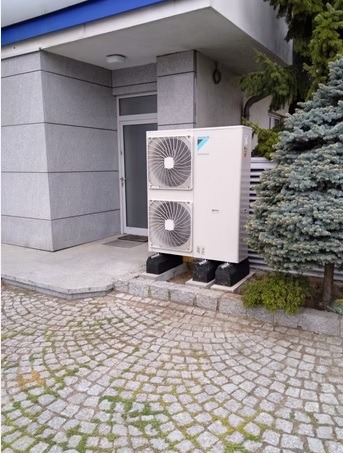 Biała pompa ciepła Daikin na czarnych podstawach, zainstalowana przy szarym budynku z drzwiami i ozdobnym brukiem. Obok rośnie iglak.