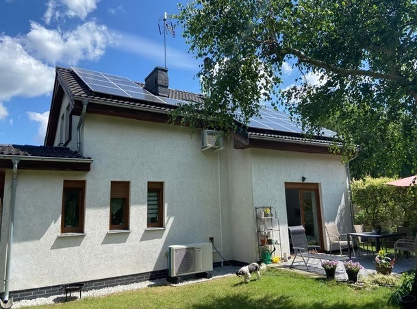 Biały dom z panelami słonecznymi na dachu i klimatyzatorem na ścianie, otoczony zielonym trawnikiem i drzewami. Pies biega po trawie, a w tle widoczny jest taras.