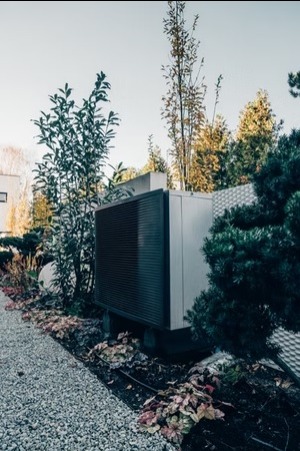 Nowoczesna pompa ciepła powietrze-woda, zainstalowana w ogrodzie z ozdobnymi roślinami i ścieżką żwirową. Widok z boku, w tle budynek mieszkalny.