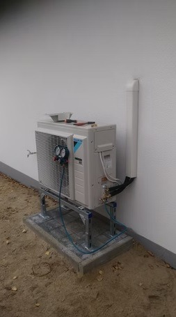 Biała zewnętrzna jednostka klimatyzacji Daikin zamontowana na metalowej konstrukcji przy ścianie budynku. Widoczne przewody i manometry.