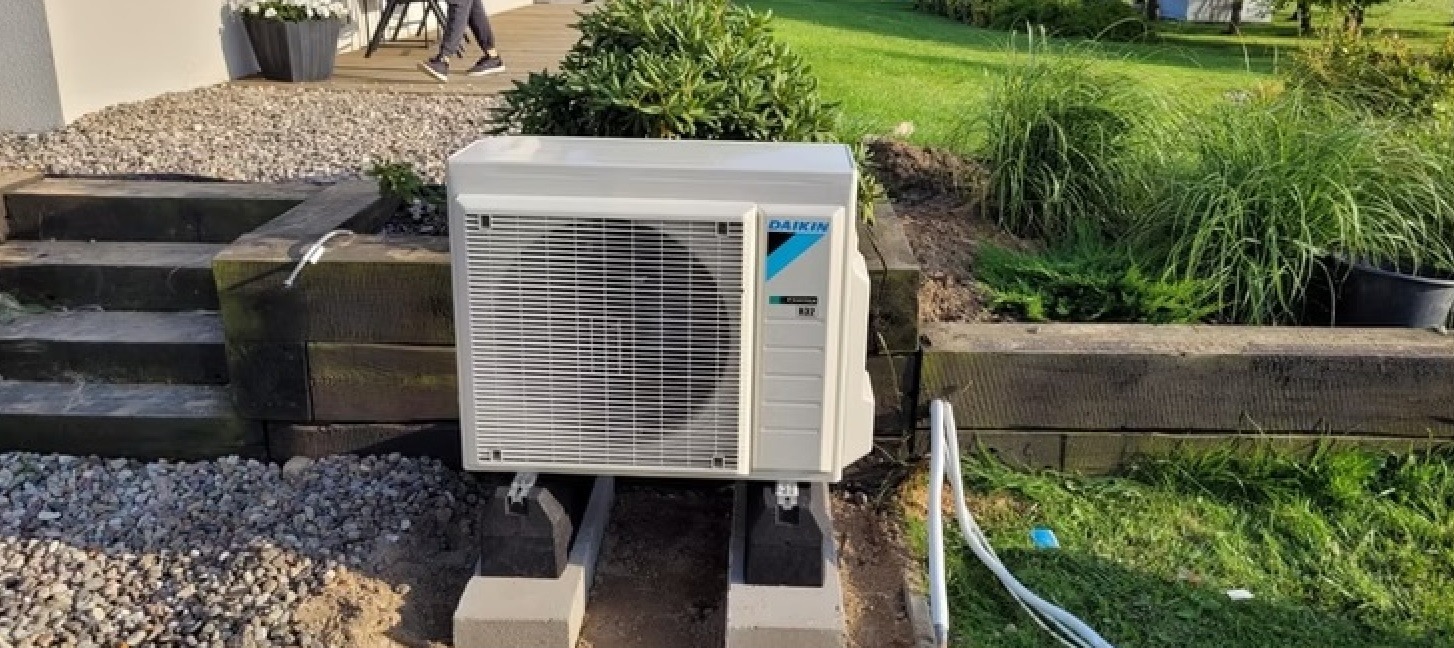 Biała jednostka zewnętrzna pompy ciepła Daikin, zamontowana na betonowych wspornikach w ogrodzie, z widocznymi przewodami i zielenią w tle. Ujęcie z poziomu gruntu.