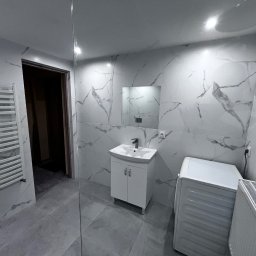 KrissGlaz - Wykończona łazienka w Pierściecu z białymi płytkami imitującymi marmur, białą szafką z umywalką i grzejnikiem drabinkowym. Widoczna pralka i lustro.