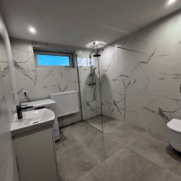 KrissGlaz - Nowoczesna łazienka z kabiną prysznicową typu walk-in, umywalką z szafką, toaletą podwieszaną i grzejnikiem drabinkowym. Białe płytki imitujące marmur z szarymi żyłami.