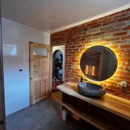 KrissGlaz - Nowoczesna łazienka z ceglaną ścianą, okrągłym lustrem z podświetleniem LED, kamienną umywalką na drewnianym blacie i drewnianymi drzwiami z szybą.