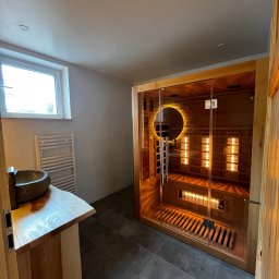 KrissGlaz - Nowoczesna łazienka z sauną infrared: kamienna umywalka na drewnianym blacie, szare płytki podłogowe, przeszklona kabina sauny z podświetleniem i drewnianym wykończeniem.