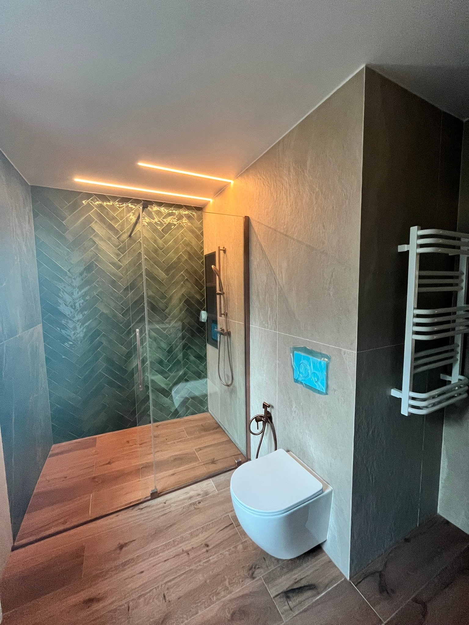 Nowoczesna łazienka z kabiną prysznicową wyłożoną zieloną jodełką, drewnopodobną podłogą i podświetleniem LED. Minimalistyczna toaleta podwieszana.