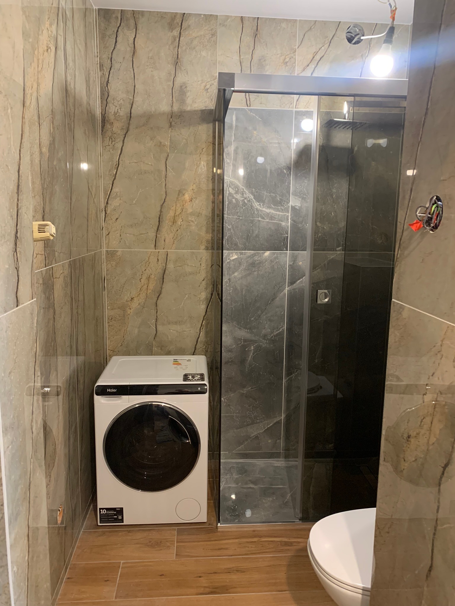 Wykończona łazienka z kabiną prysznicową typu walk-in, pralką i toaletą. Płytki imitujące marmur na ścianach i podłodze. Widoczne niedokończone podłączenie oświetlenia.