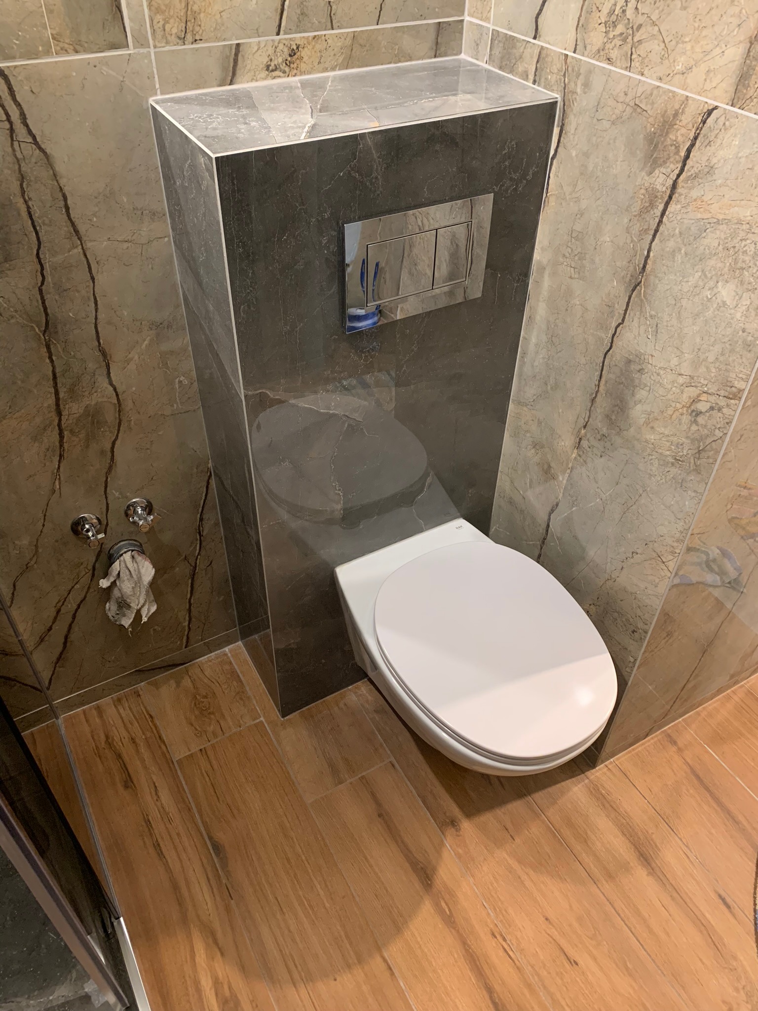 Nowoczesna toaleta podwieszana z szarą obudową i chromowanym przyciskiem spłukującym, otoczona marmurowymi płytkami w odcieniach beżu i brązu. Widoczne drewnopodobne płytki podłogowe.