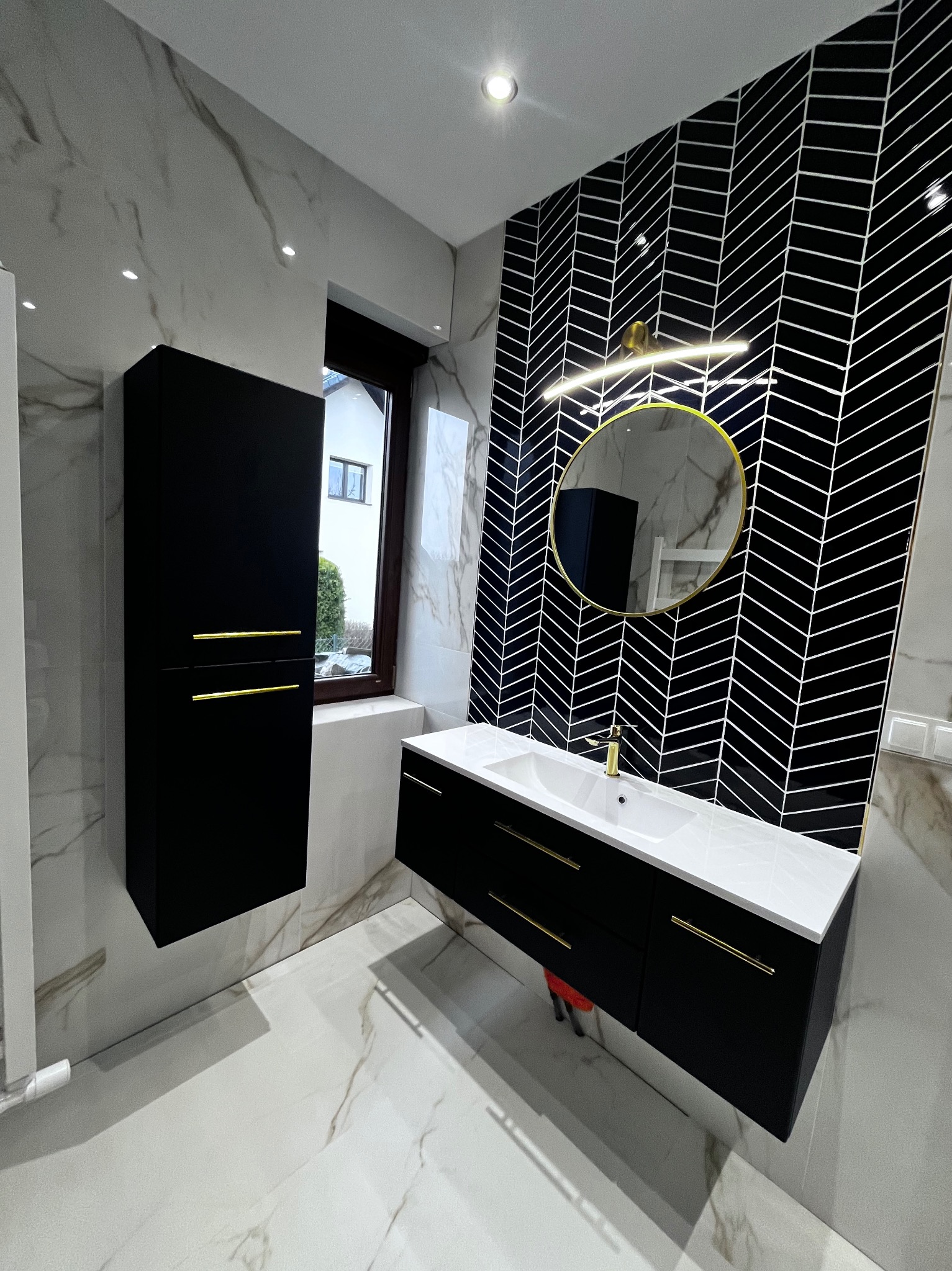 Nowoczesna łazienka z czarno-białymi płytkami ułożonymi w jodełkę, czarnymi szafkami z złotymi uchwytami i okrągłym lustrem w złotej ramie. Marmurowe płytki na ścianach i podłodze.