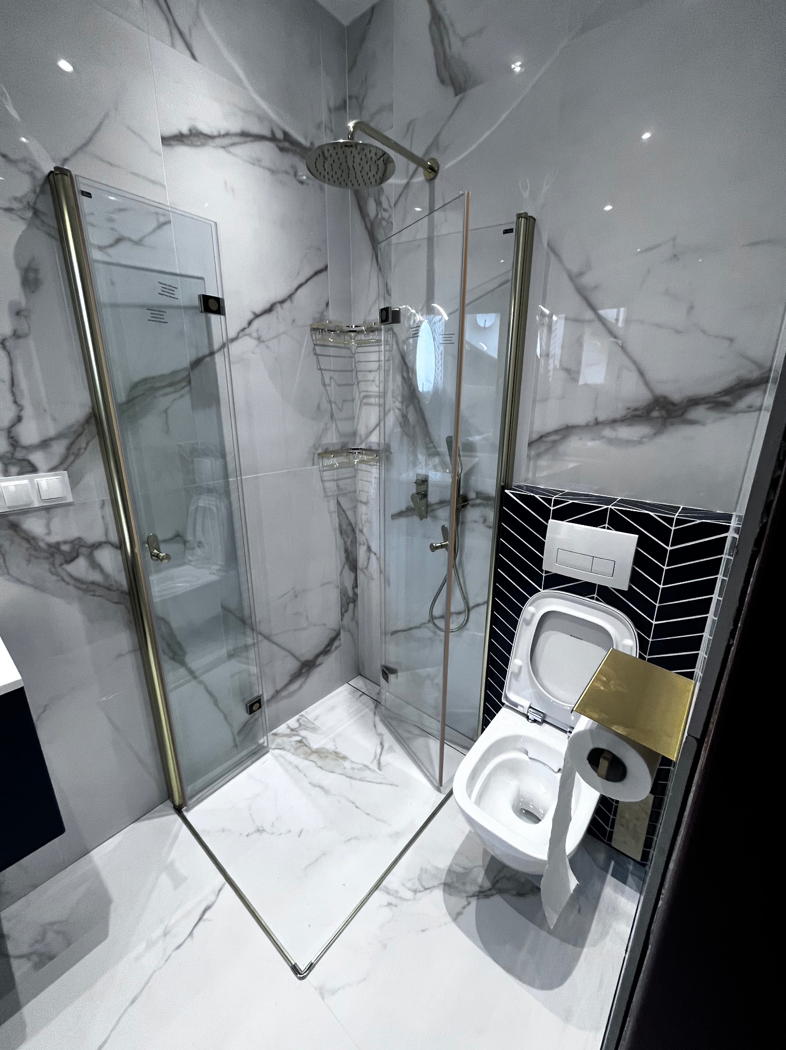 Nowoczesna łazienka z kabiną prysznicową typu walk-in, białe płytki imitujące marmur i toaleta z czarną ścianą w geometryczny wzór. Chromowana armatura.