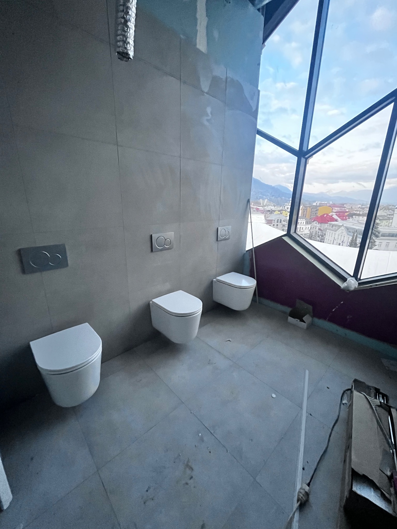 Montaż podtynkowych toalet w łazience z dużym oknem i widokiem na miasto. Szare płytki na ścianach i podłodze. Widoczne elementy instalacji i narzędzia.