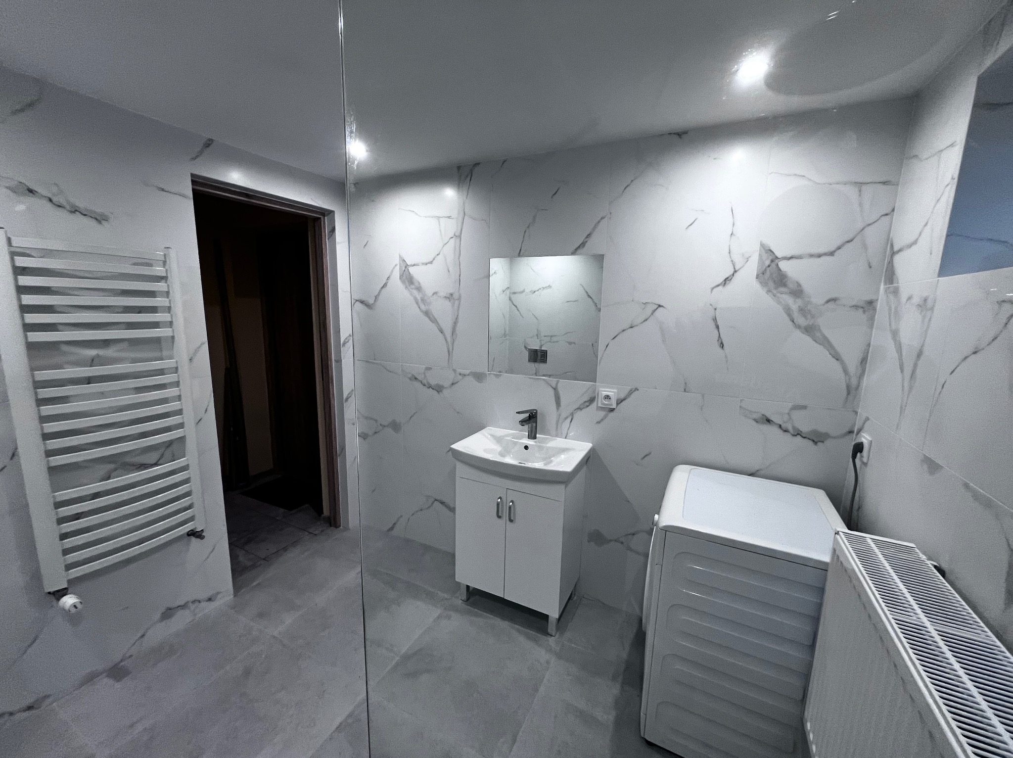 Wykończona łazienka w Pierściecu z białymi płytkami imitującymi marmur, białą szafką z umywalką i grzejnikiem drabinkowym. Widoczna pralka i lustro.