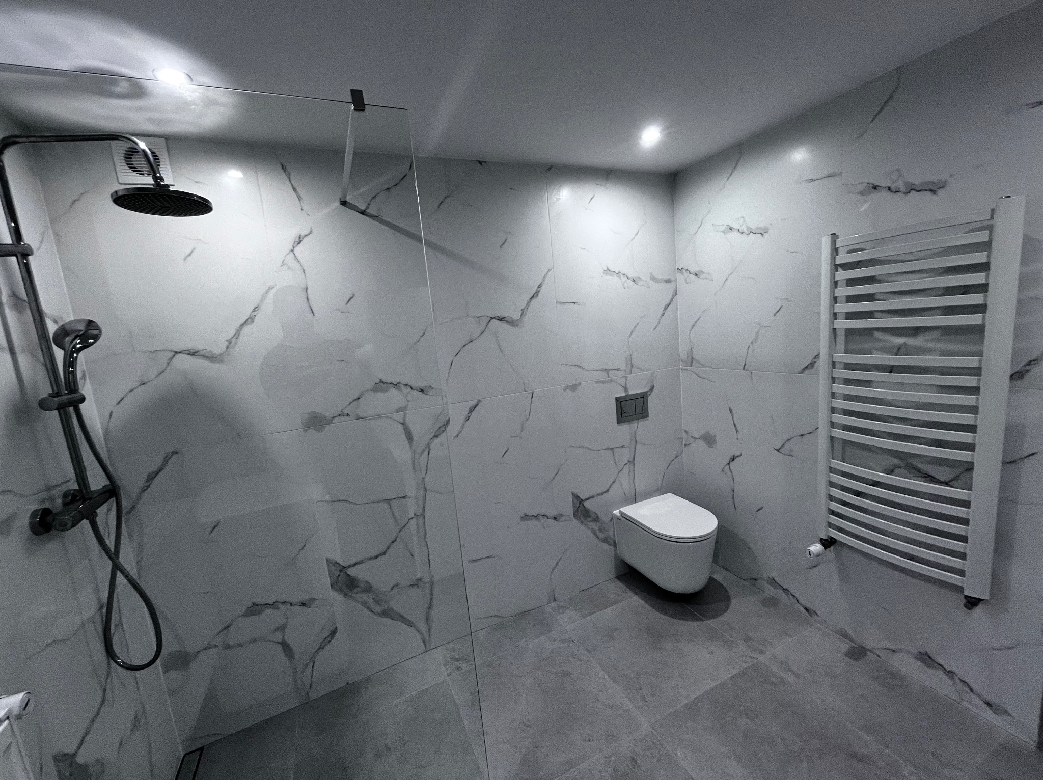 Nowoczesna łazienka z marmurowymi płytkami, kabiną prysznicową typu walk-in, podwieszaną toaletą i grzejnikiem drabinkowym. Minimalistyczny design, szaro-biała kolorystyka.