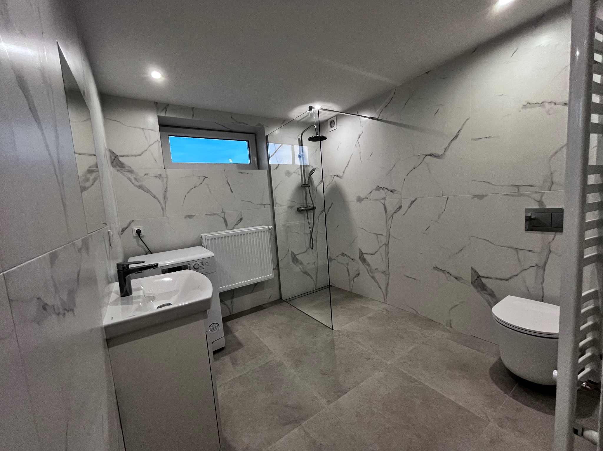 Nowoczesna łazienka z kabiną prysznicową typu walk-in, umywalką z szafką, toaletą podwieszaną i grzejnikiem drabinkowym. Białe płytki imitujące marmur z szarymi żyłami.