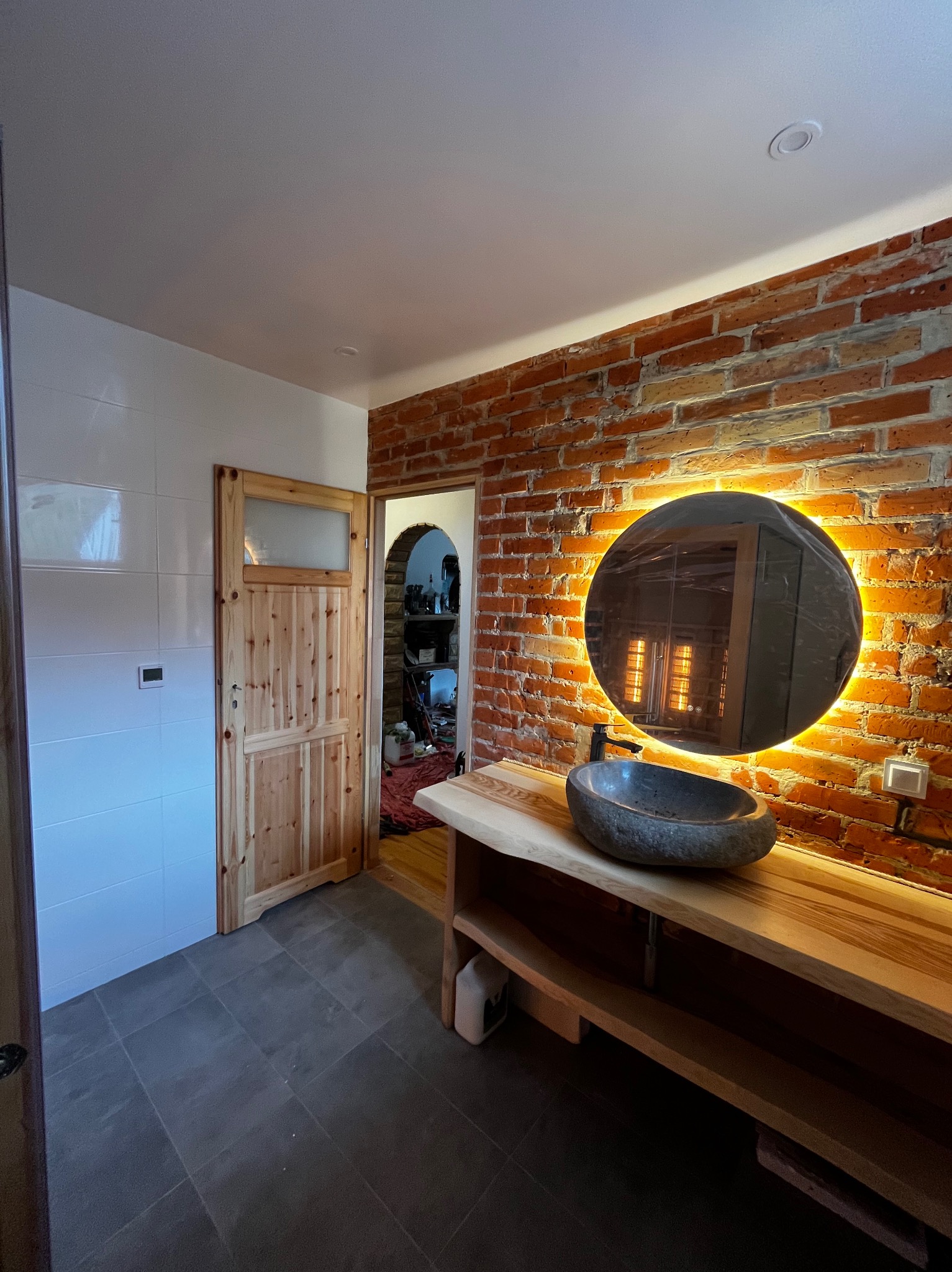 Nowoczesna łazienka z ceglaną ścianą, okrągłym lustrem z podświetleniem LED, kamienną umywalką na drewnianym blacie i drewnianymi drzwiami z szybą.