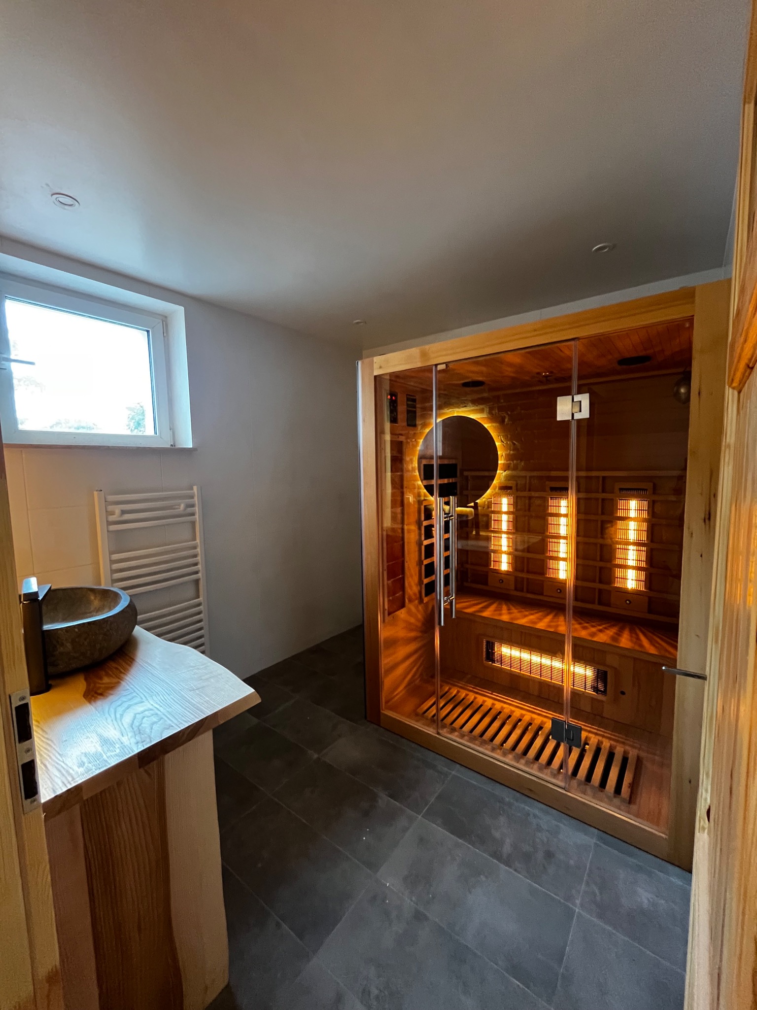 Nowoczesna łazienka z sauną infrared: kamienna umywalka na drewnianym blacie, szare płytki podłogowe, przeszklona kabina sauny z podświetleniem i drewnianym wykończeniem.