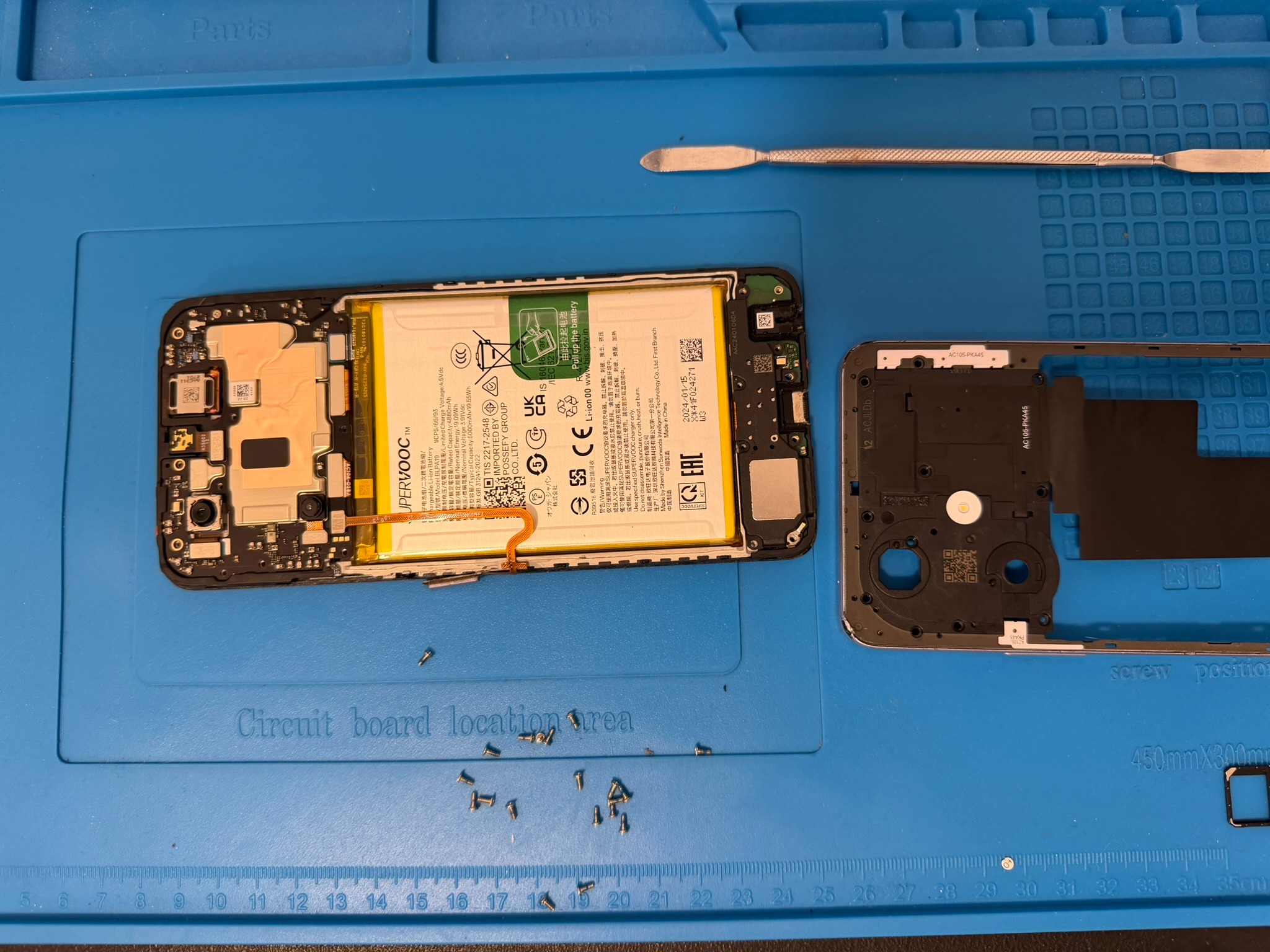 Rozłożony smartfon z widoczną baterią, narzędziem i śrubkami na macie serwisowej. Wymiana baterii w serwisie telefonów, widok z góry.