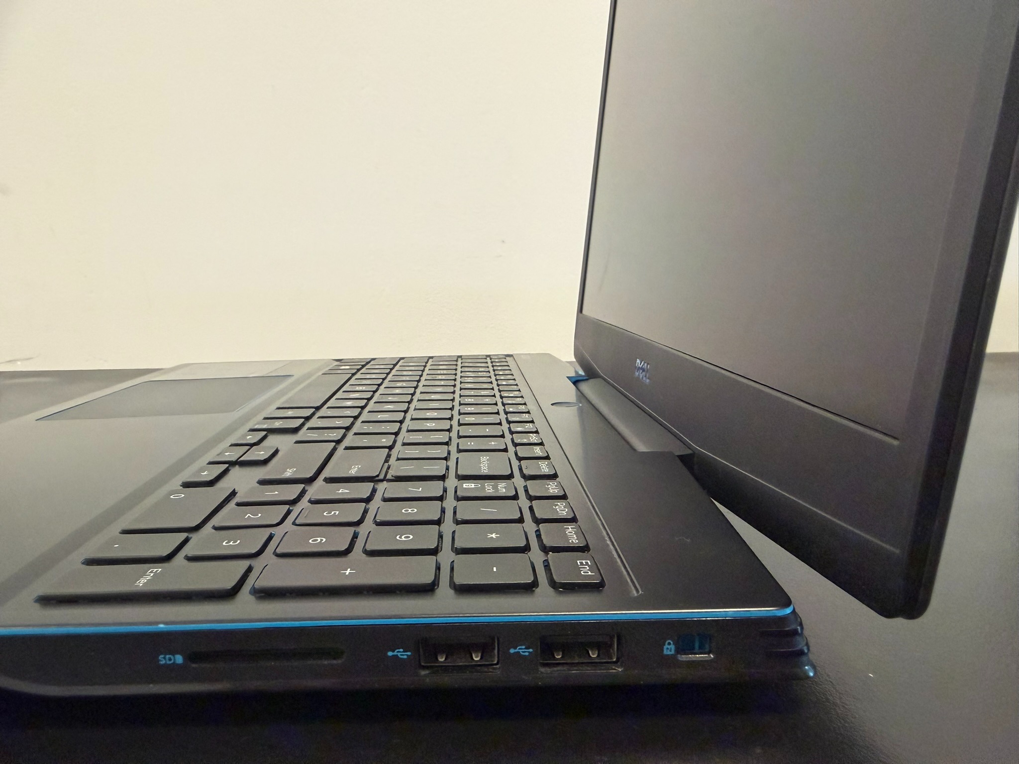 Czarny laptop Dell z widoczną klawiaturą, portami USB i czytnikiem kart SD, ustawiony na ciemnej powierzchni, z białym tłem. Widok z boku, lekko z dołu.