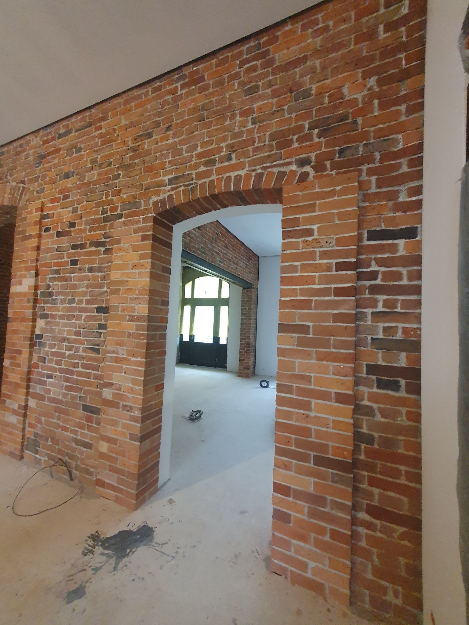 Ceglana ściana z łukowym przejściem do jasnego pomieszczenia. Widoczne detale faktury cegieł i surowy charakter wnętrza w trakcie prac wykończeniowych.