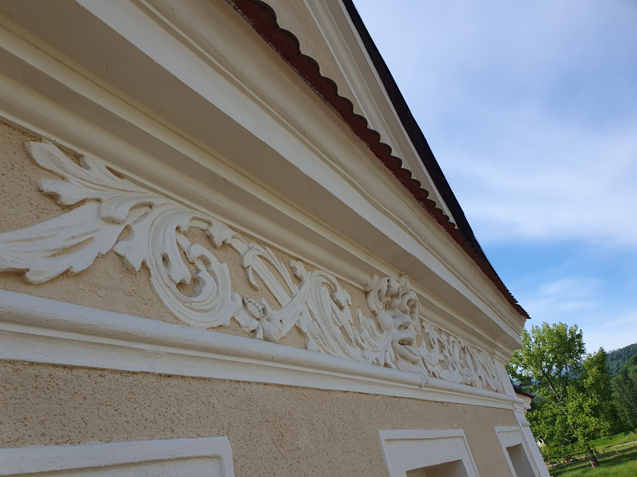 Detal fasady budynku z dekoracyjnym, białym ornamentem sztukatorskim na tle beżowej ściany i błękitnego nieba. Widoczny fragment dachu i drzewo w tle.