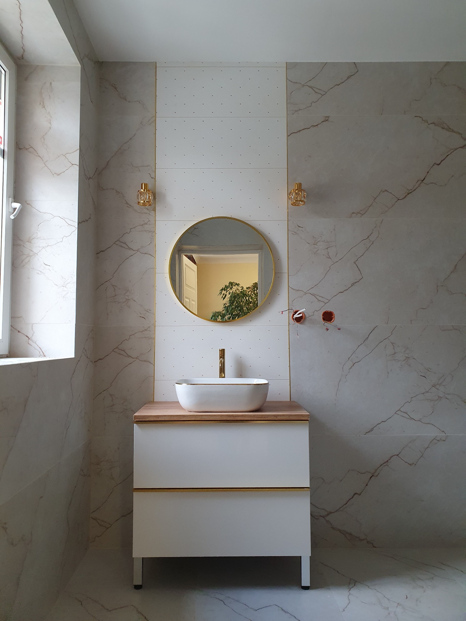 Elegancka łazienka z marmurowymi płytkami, okrągłym lustrem w złotej ramie i białą szafką pod umywalkę z drewnianym blatem. Widoczne kinkiety i fragment instalacji.