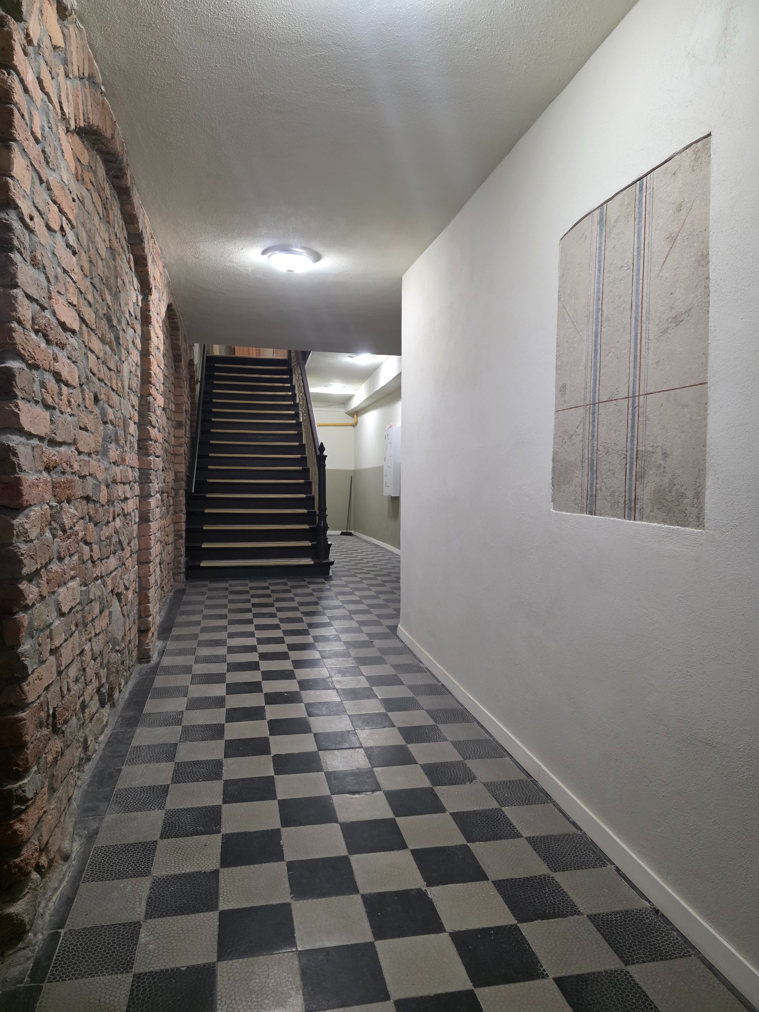 Korytarz z ceglaną ścianą, schodami i wzorzystą podłogą w szachownicę. Na ścianie widoczny fragment odsłoniętej warstwy z pionowymi liniami. Oświetlenie sufitowe.