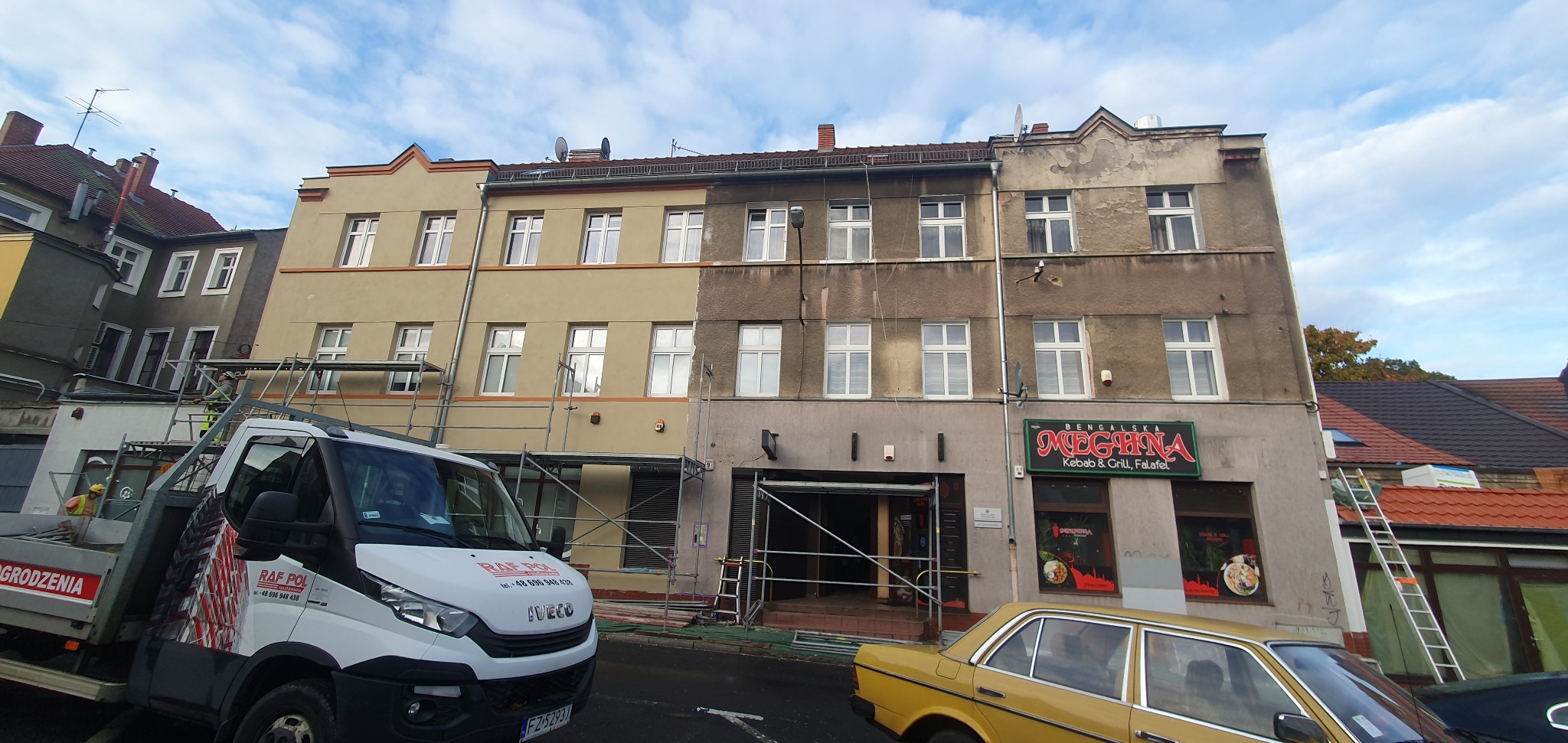 Budynek z elewacją w trakcie odnawiania: połowa pomalowana na beżowo, druga w stanie surowym. Widoczne rusztowania, samochód dostawczy i żółty samochód osobowy.