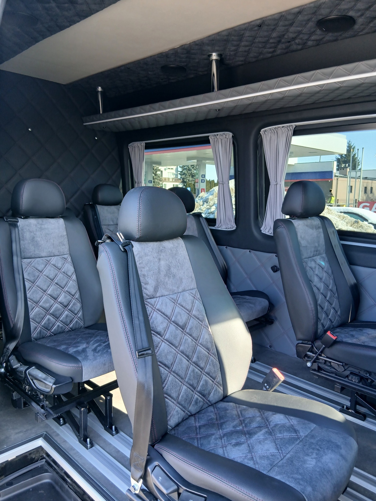 Wnętrze luksusowego busa z czarną, pikowaną tapicerką, szarymi zasłonkami w oknach i regulowanymi fotelami. Widok na stację benzynową za oknem.