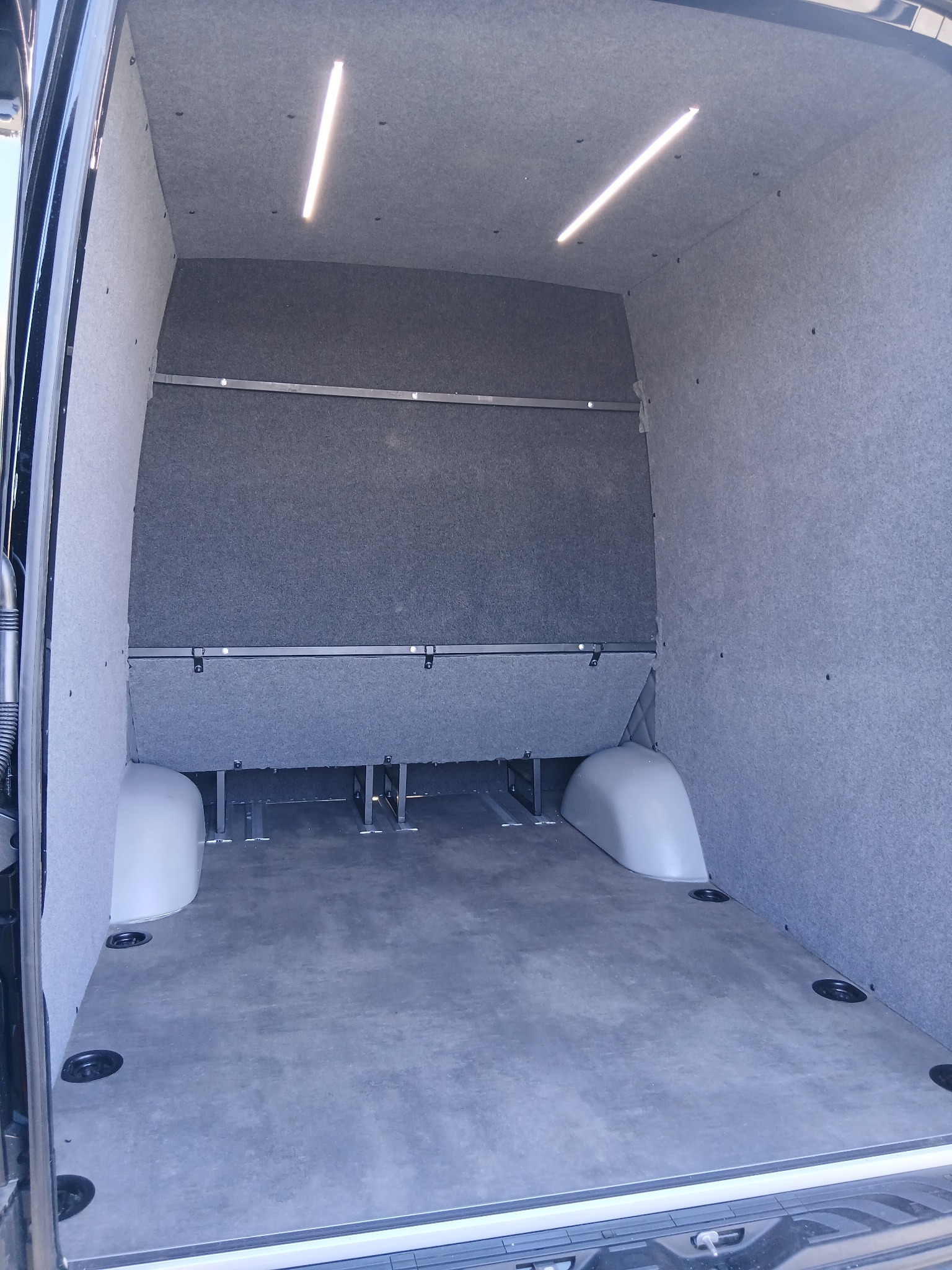 Wnętrze wykończonego busa z szarą tapicerką ścian i sufitu, oświetleniem LED i zamontowanymi szynami mocującymi. Widok z tyłu pojazdu na pustą przestrzeń ładunkową.