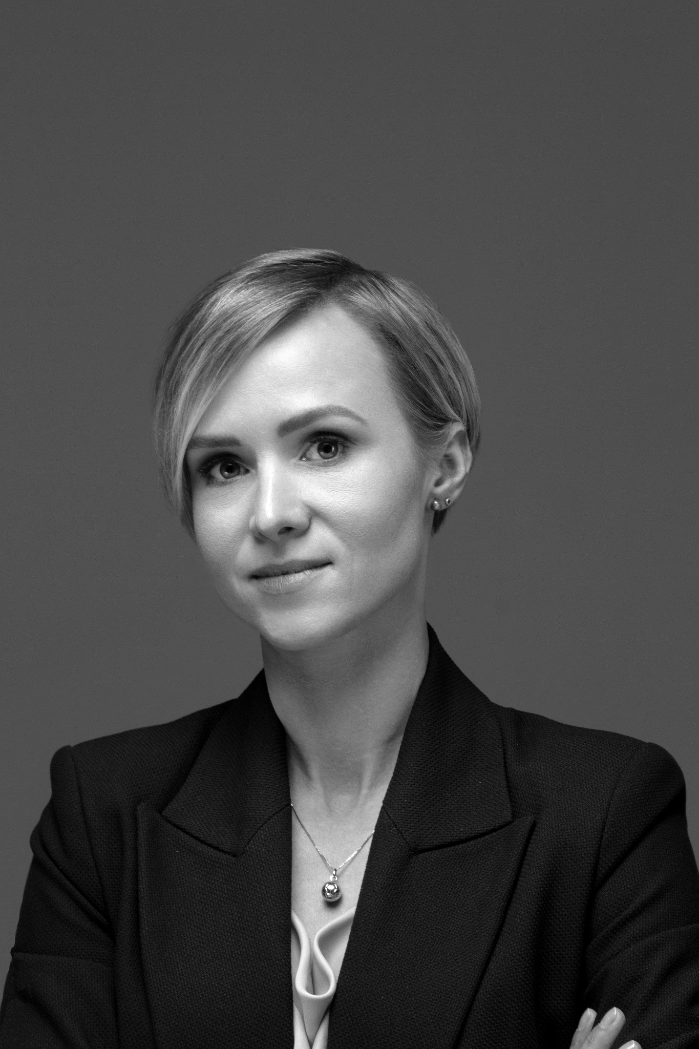 Portret kobiety w czarnym żakiecie na szarym tle. Krótkie blond włosy, delikatny makijaż, srebrny naszyjnik. Profesjonalny wizerunek.