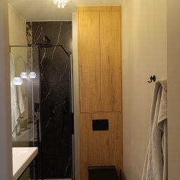 Wojciech Ulrych - Nowoczesna łazienka z drewnianą szafką w zabudowie, czarną armaturą i kabiną prysznicową z marmurowym wykończeniem. Kryształowy żyrandol dodaje elegancji.