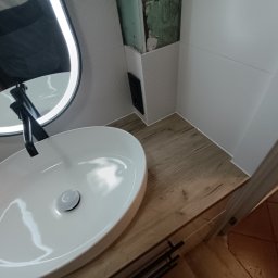 Wojciech Ulrych - Nowoczesna łazienka z owalną umywalką nablatową, czarną armaturą i podświetlanym lustrem. Widoczne białe płytki i drewnopodobny blat szafki. Fragment ściany w trakcie wykończenia.