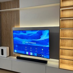 Wojciech Ulrych - Nowoczesna zabudowa ściany z TV, z drewnianymi lamelami i podświetlanymi półkami. Minimalistyczny design, białe szafki dolne i szary panel za telewizorem.