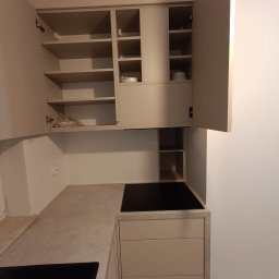 Wojciech Ulrych - Jasna kuchnia z otwartymi szafkami, widoczna płyta indukcyjna i blat o teksturze betonu. Minimalistyczny design, ciepłe oświetlenie.