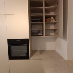 Wojciech Ulrych - Jasna kuchnia na wymiar z zabudowaną piekarnikiem i otwartą szafką z półkami. Widoczne przedmioty wewnątrz szafki. Minimalistyczny design.