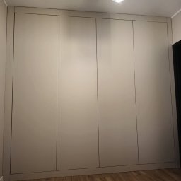 Czterodrzwiowa szafa wnękowa w odcieniach beżu, z minimalistycznymi uchwytami, dopasowana do wnętrza z drewnianą podłogą i nowoczesnym oświetleniem sufitowym.