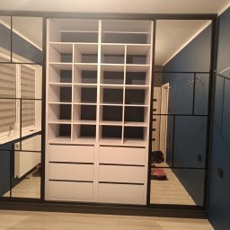 Biała szafa wnękowa z czarnymi ramami i lustrami w Barcinie. Widoczne półki i szuflady. Nowoczesny design, niebieska ściana w tle.