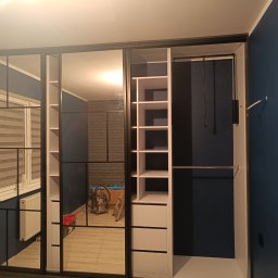 Nowoczesna szafa wnękowa z przesuwnymi drzwiami lustrzanymi i czarnymi ramami, wnętrze z półkami i szufladami, na tle granatowej ściany.