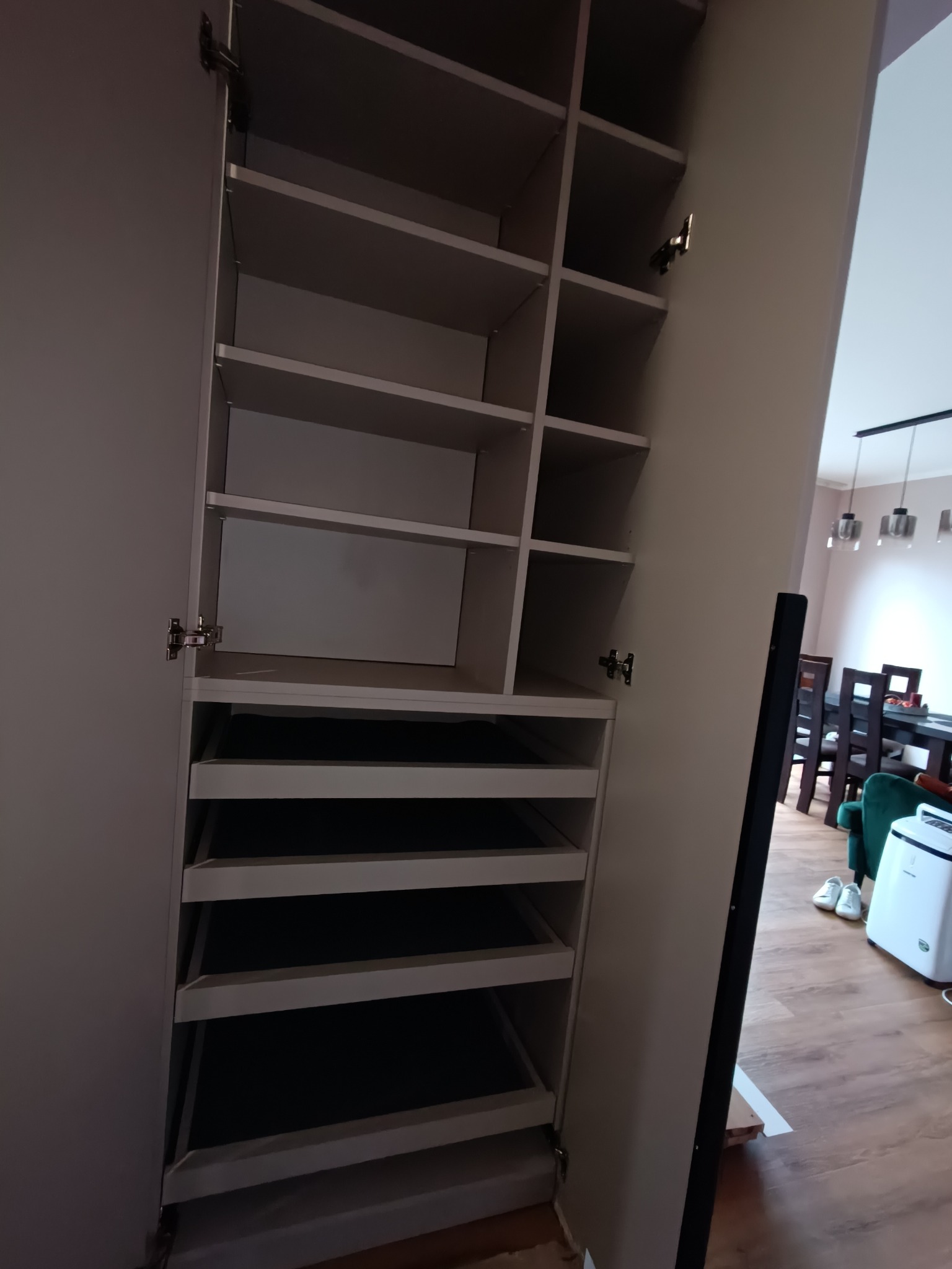 Otwarta, jasna szafa wnękowa z półkami i szufladami, widoczna wnęka i fragment jadalni w tle. Minimalistyczny design, neutralna kolorystyka.