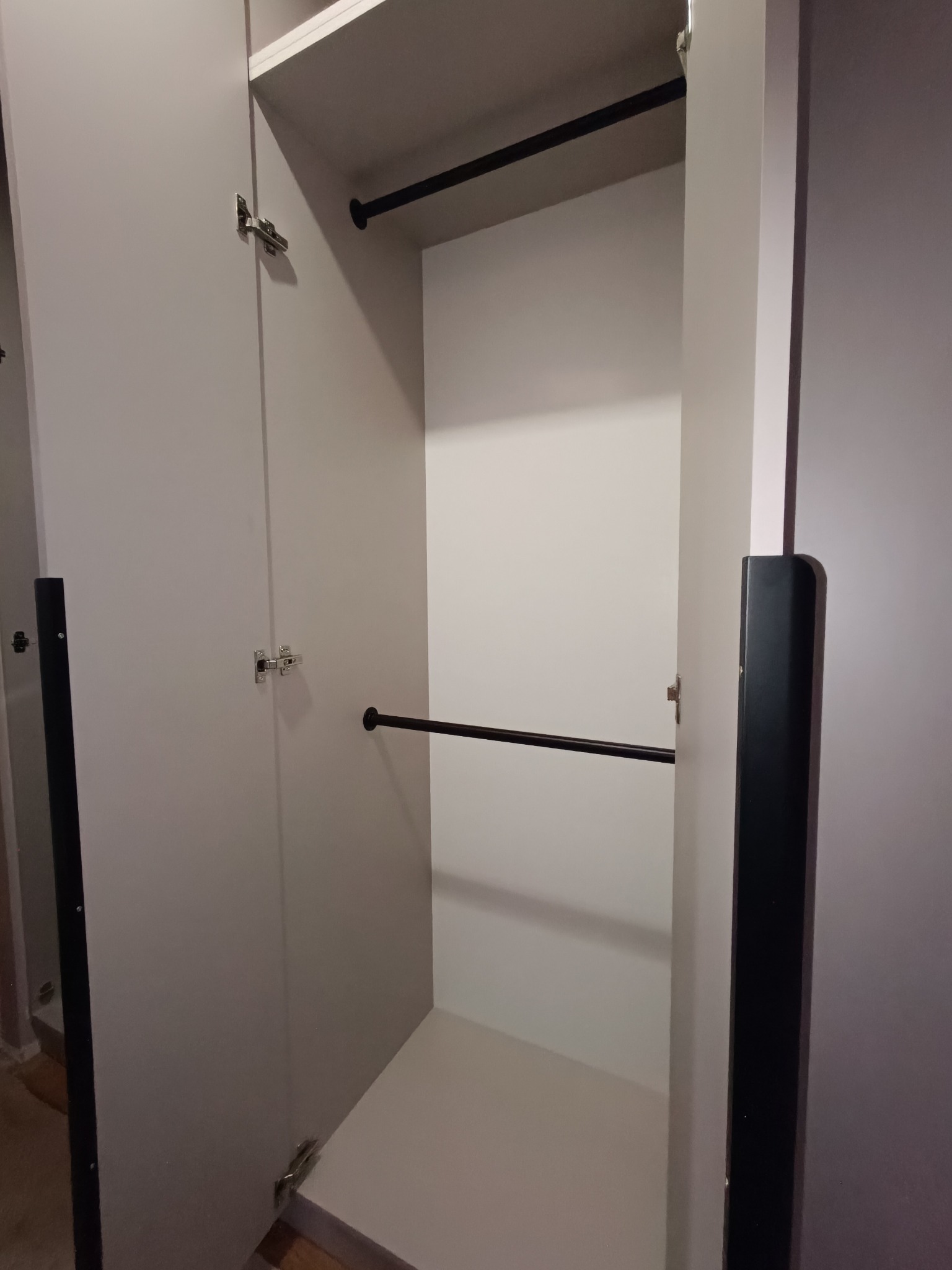 Otwarta, jasna szafa z dwoma drążkami na ubrania, widoczne zawiasy i czarne listwy boczne. Minimalistyczny design, wnętrze szafy w bieli.