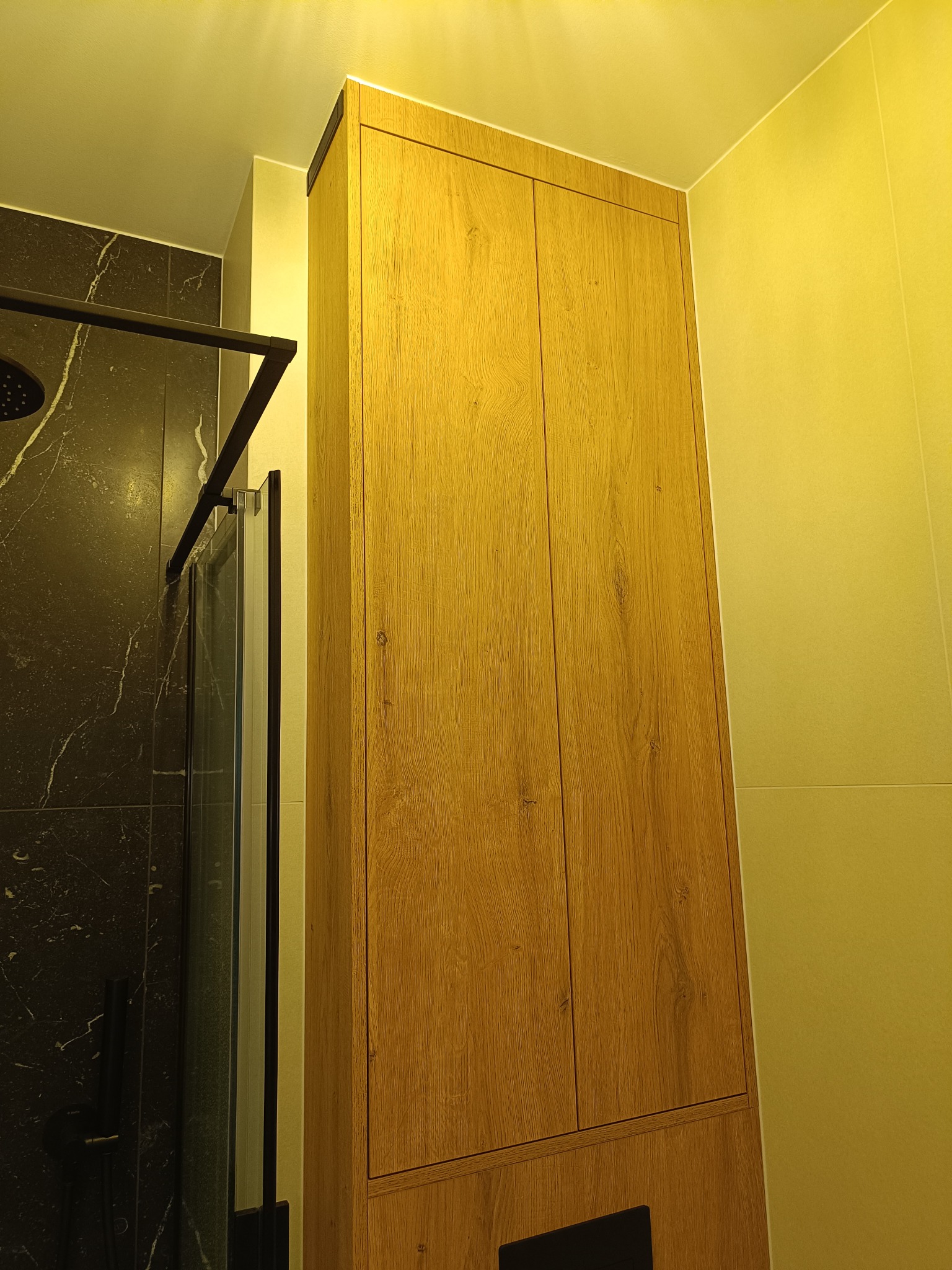 Wysoka, drewniana szafa w łazience z czarnymi elementami kabiny prysznicowej i marmurowymi płytkami. Ciepłe oświetlenie podkreśla fakturę drewna.