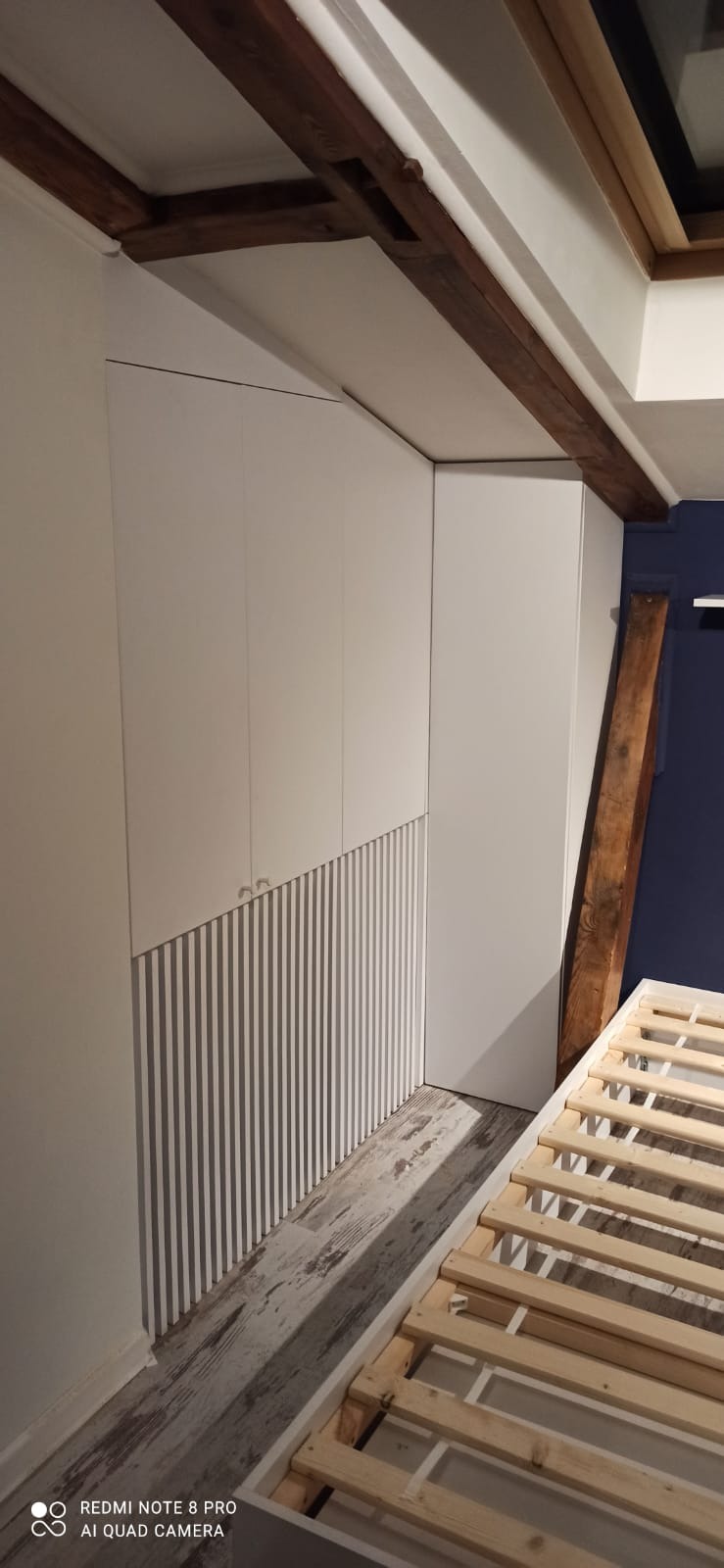 Biała szafa wnękowa z pionowymi żłobieniami, dopasowana do drewnianych belek stropowych w pokoju z surowym łóżkiem. Minimalistyczny design.