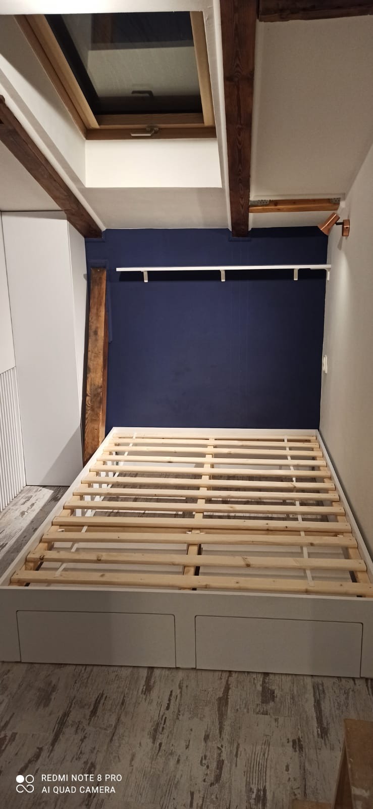 Rama łóżka z szufladami w pokoju z granatową ścianą i drewnianymi belkami stropowymi. Widoczna szafa wnękowa z białymi frontami.
