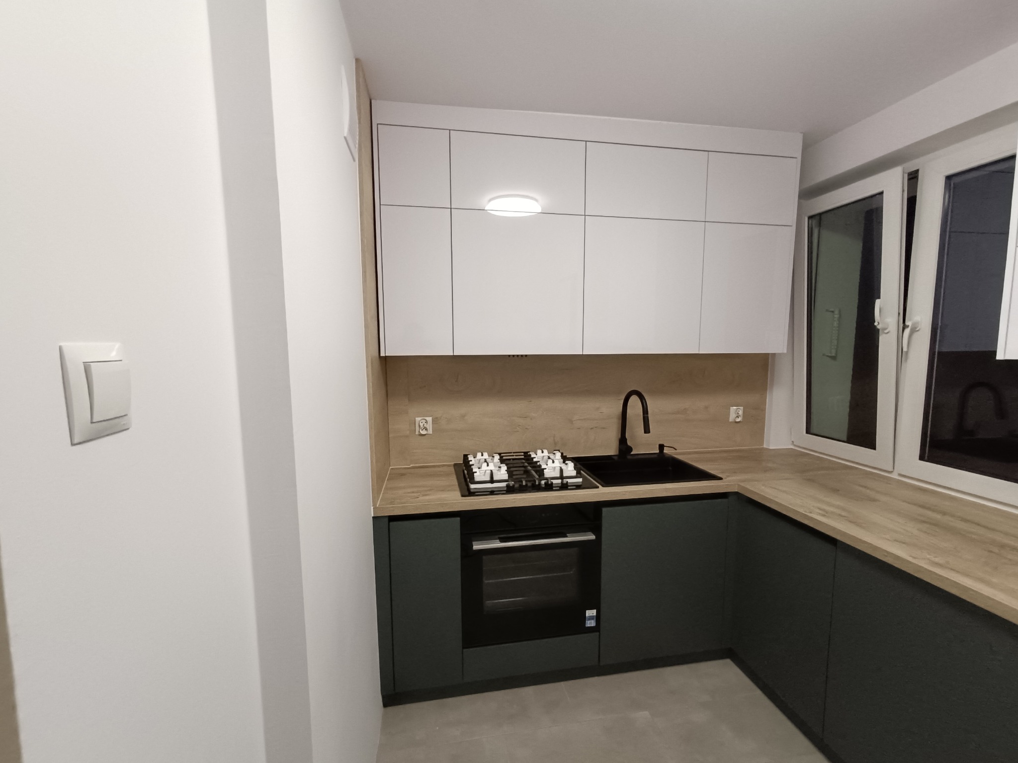 Nowoczesna kuchnia w odcieniach bieli i grafitu z drewnianym blatem, oświetleniem podszafkowym i czarną armaturą, widziana z perspektywy narożnej.