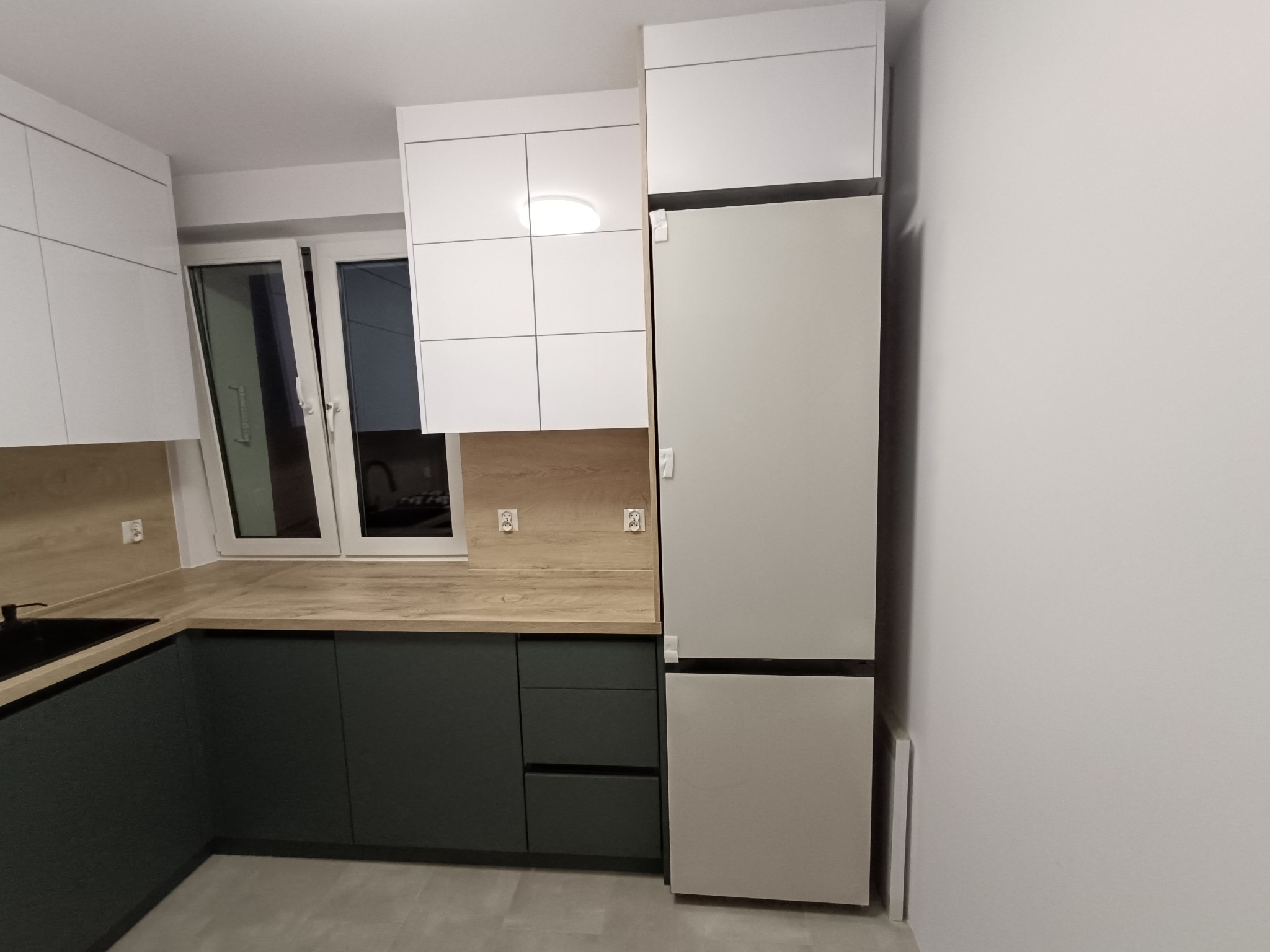 Nowoczesna kuchnia z białymi szafkami górnymi i ciemnozielonymi dolnymi, drewnianym blatem i szarą lodówką. Minimalistyczny design, oświetlenie sufitowe.