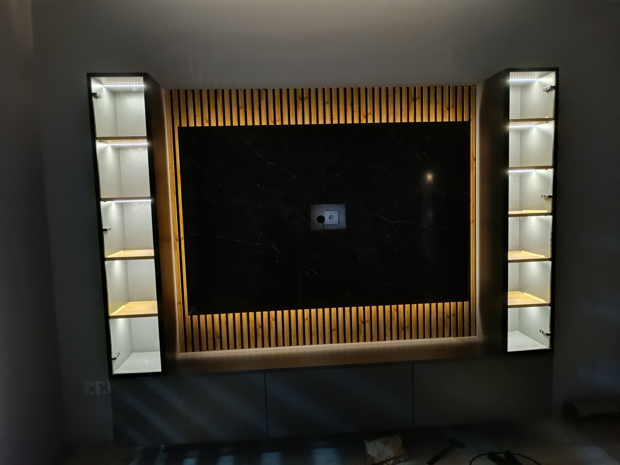 Nowoczesna zabudowa ściany z czarnym panelem imitującym marmur, drewnianymi lamelami i podświetlanymi szafkami po bokach. Minimalistyczny design z ukrytym oświetleniem.