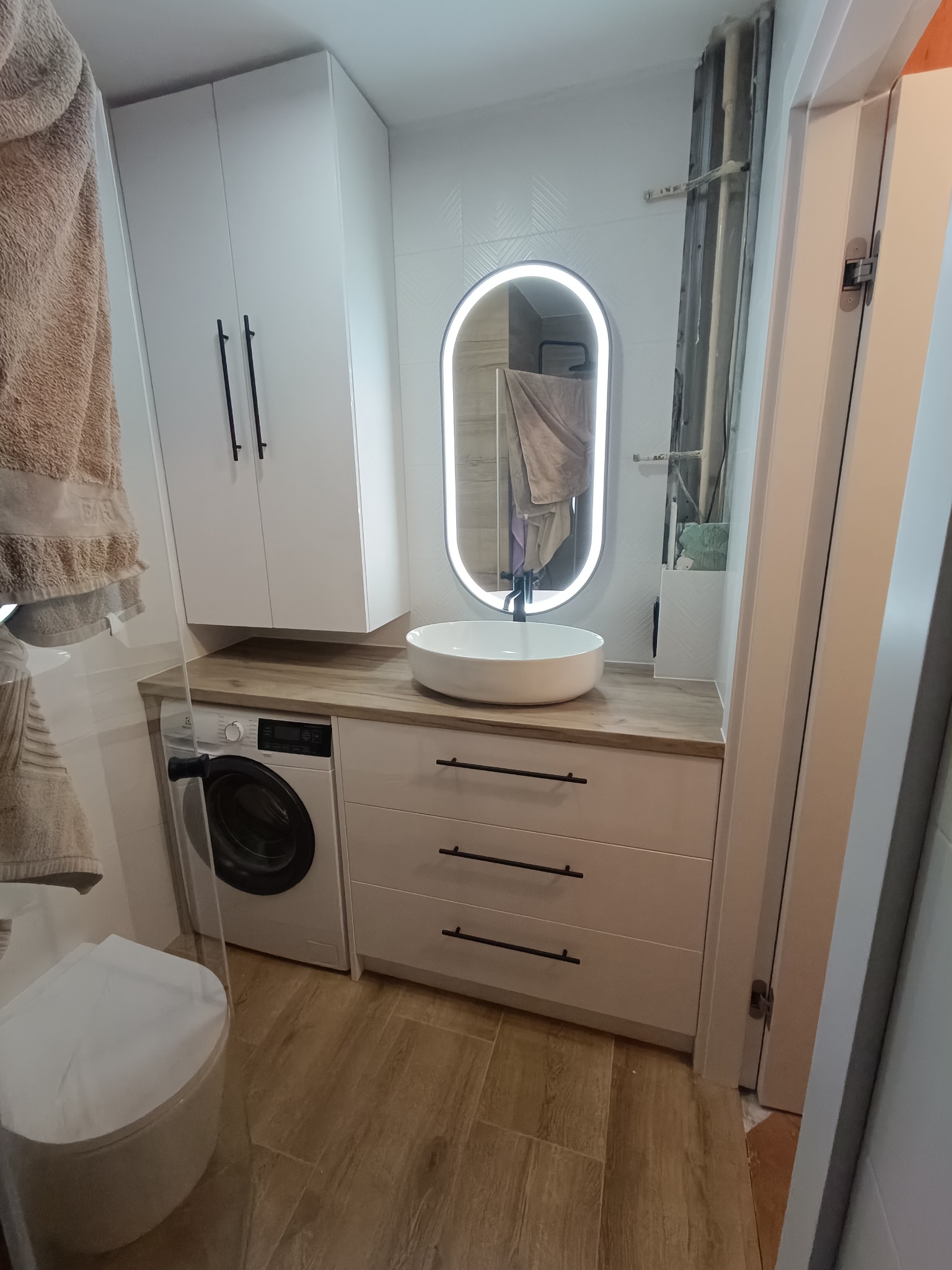 Nowoczesna łazienka z białą zabudową meblową, umywalką nablatową i podświetlanym lustrem. Obok szafki pralka, całość na tle beżowych płytek podłogowych.