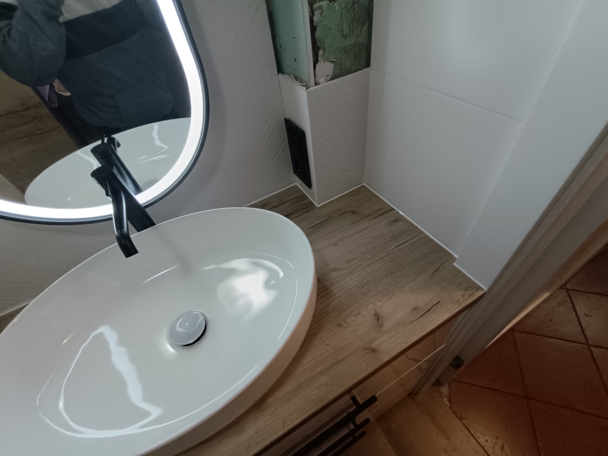 Nowoczesna łazienka z owalną umywalką nablatową, czarną armaturą i podświetlanym lustrem. Widoczne białe płytki i drewnopodobny blat szafki. Fragment ściany w trakcie wykończenia.