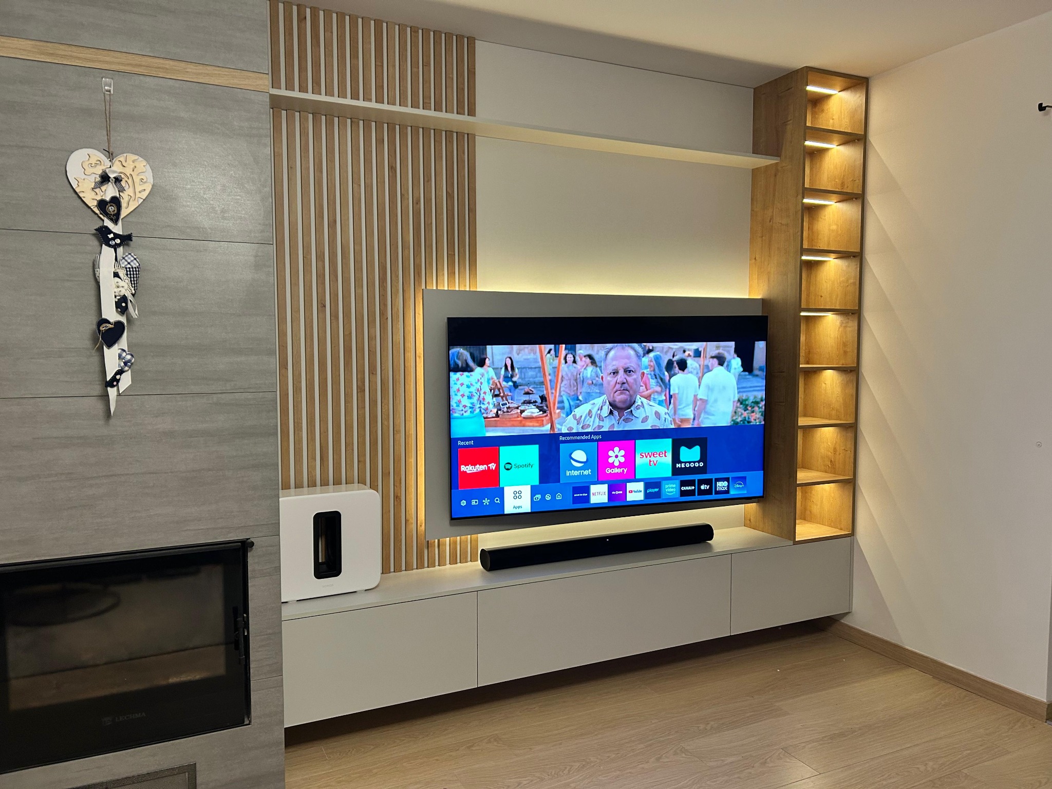 Nowoczesna zabudowa ściany z telewizorem, drewnianymi lamelami i podświetlanymi półkami. Białe szafki RTV i kominek tworzą spójną całość.